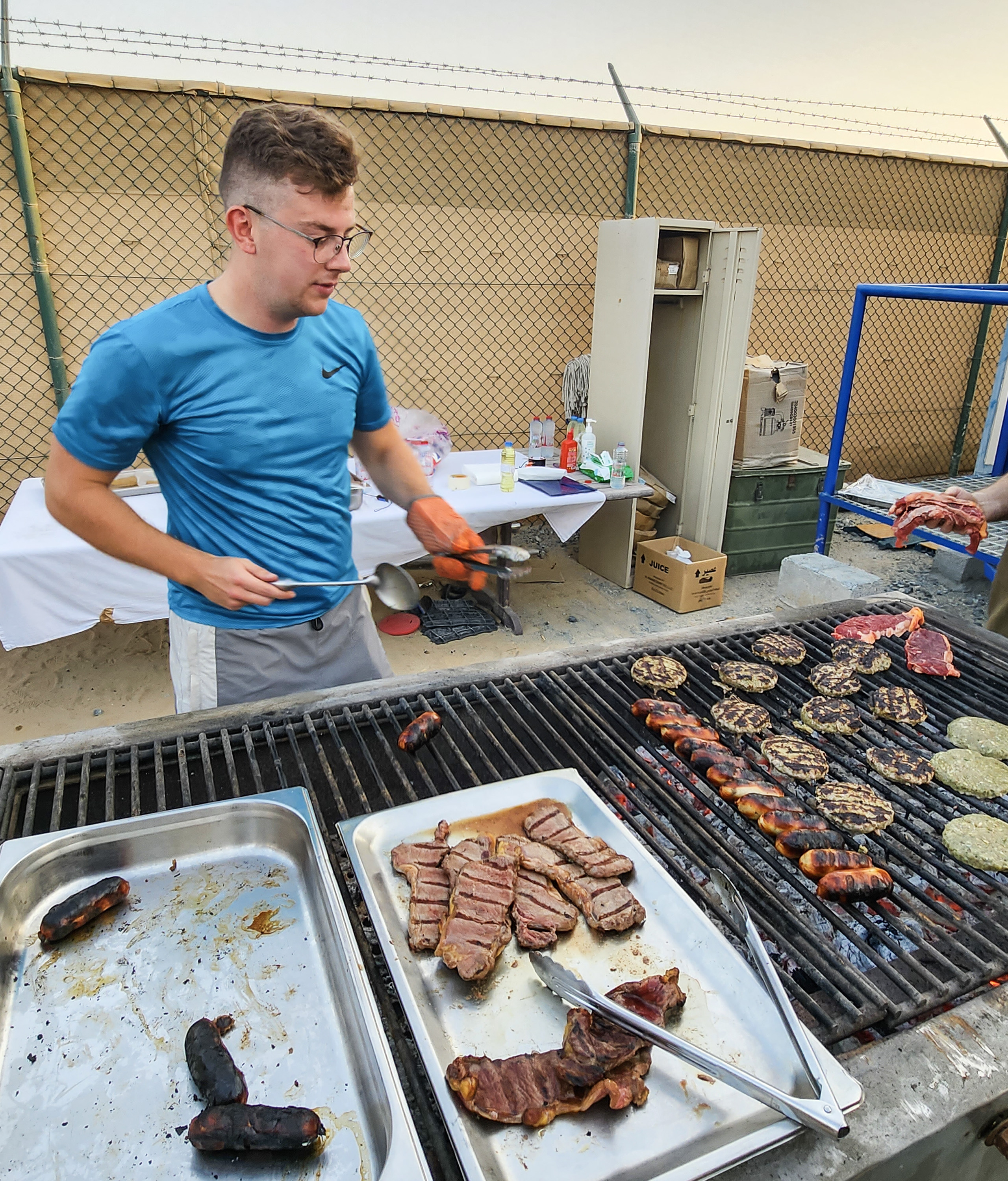 Image shows civilian at a barbecue grill cooking food.. 